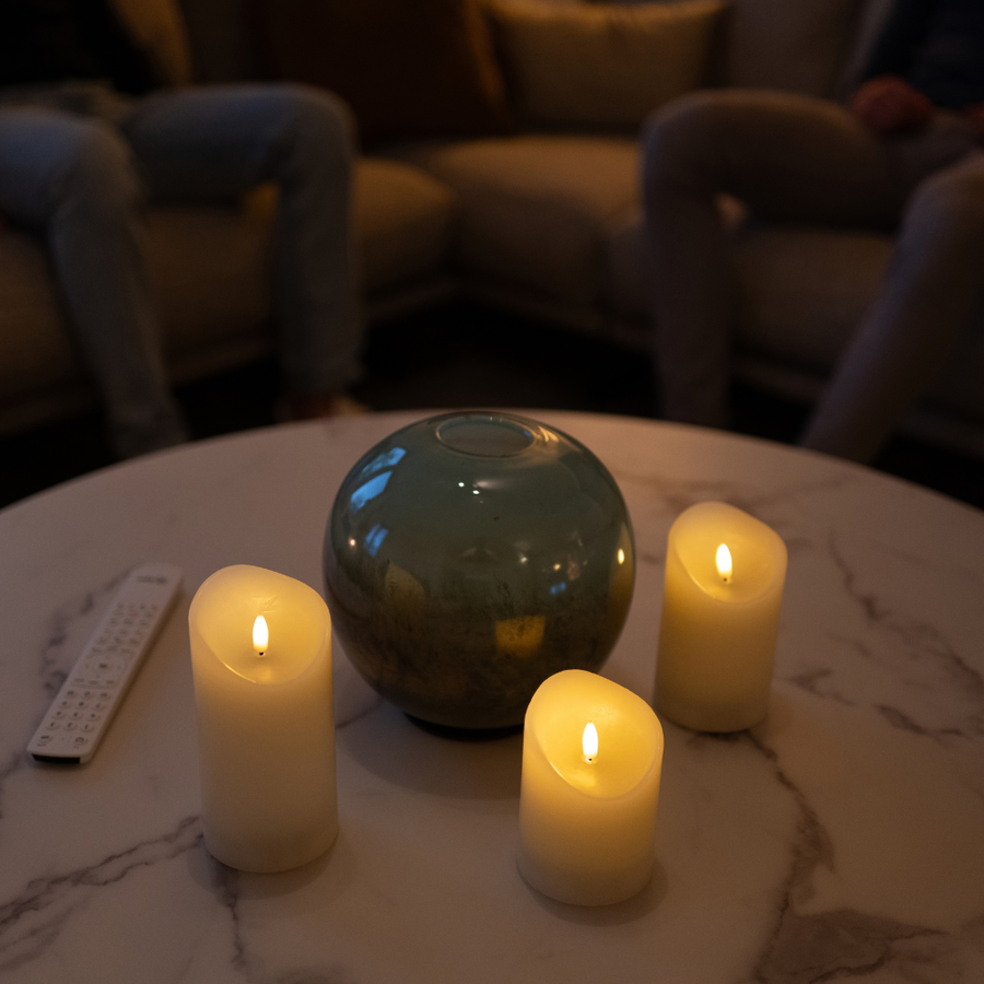 Drie Akaro kaarsen en de afstandsbediening op een marmeren salontafel, mensen op de bank erachter