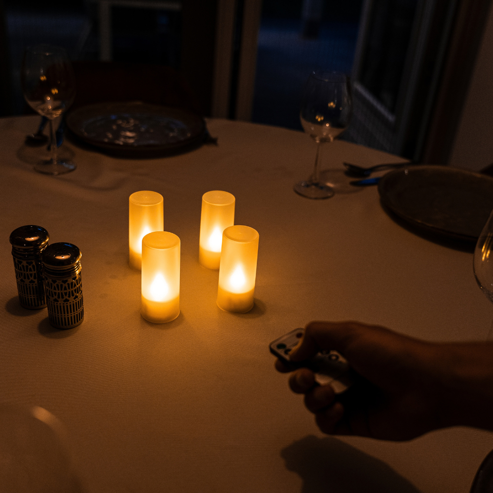 Een hand bedient met de afstandsbediening vier Akaro waxinelichtjes op een gedekte tafel